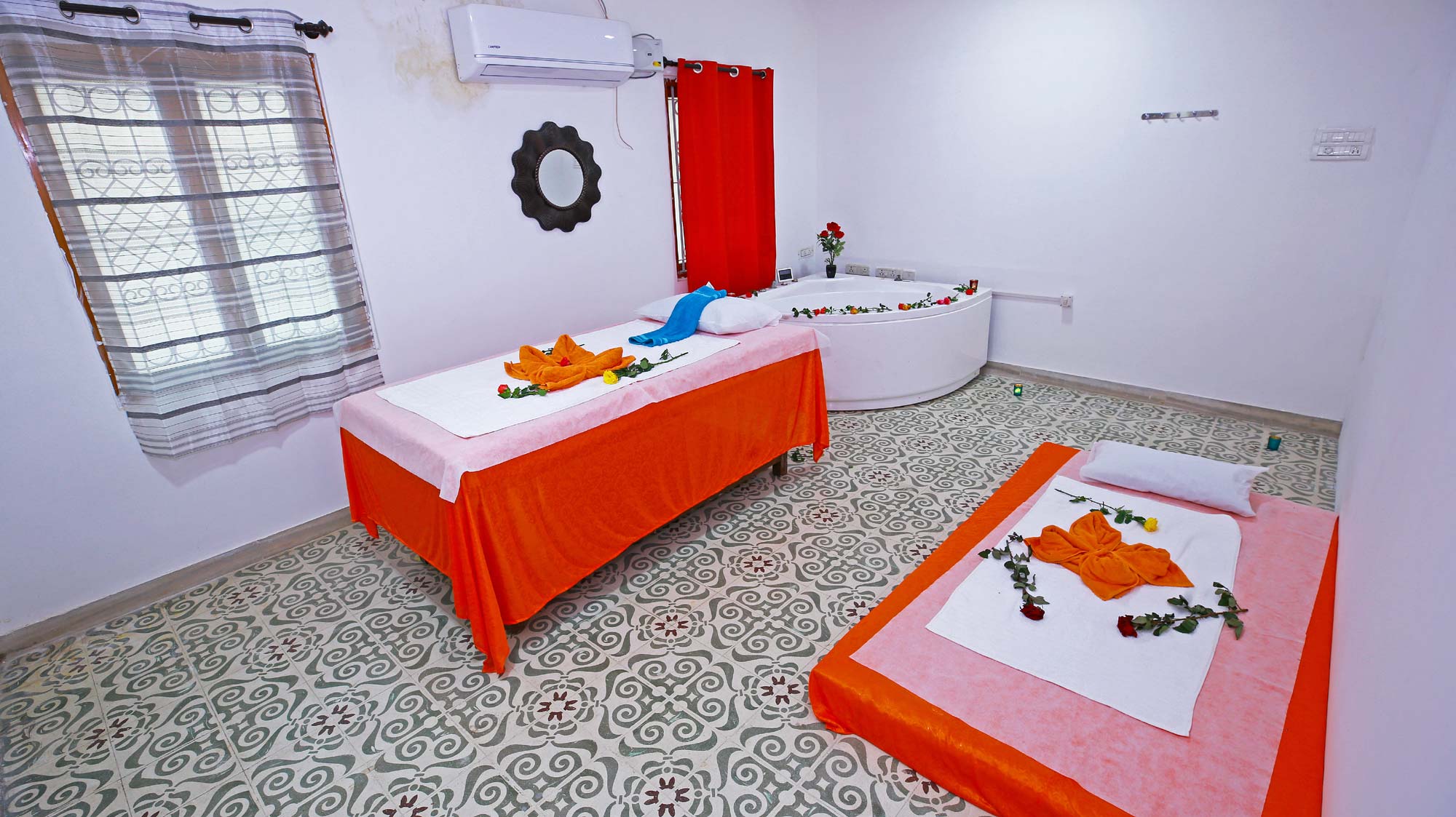 A room in a massage spa features a bathtub, massage table with pillow, towel and flowers laid on, bed with towel & flowers laid on, Air Conditioner, two windows with curtains, mirror and a hanger.
