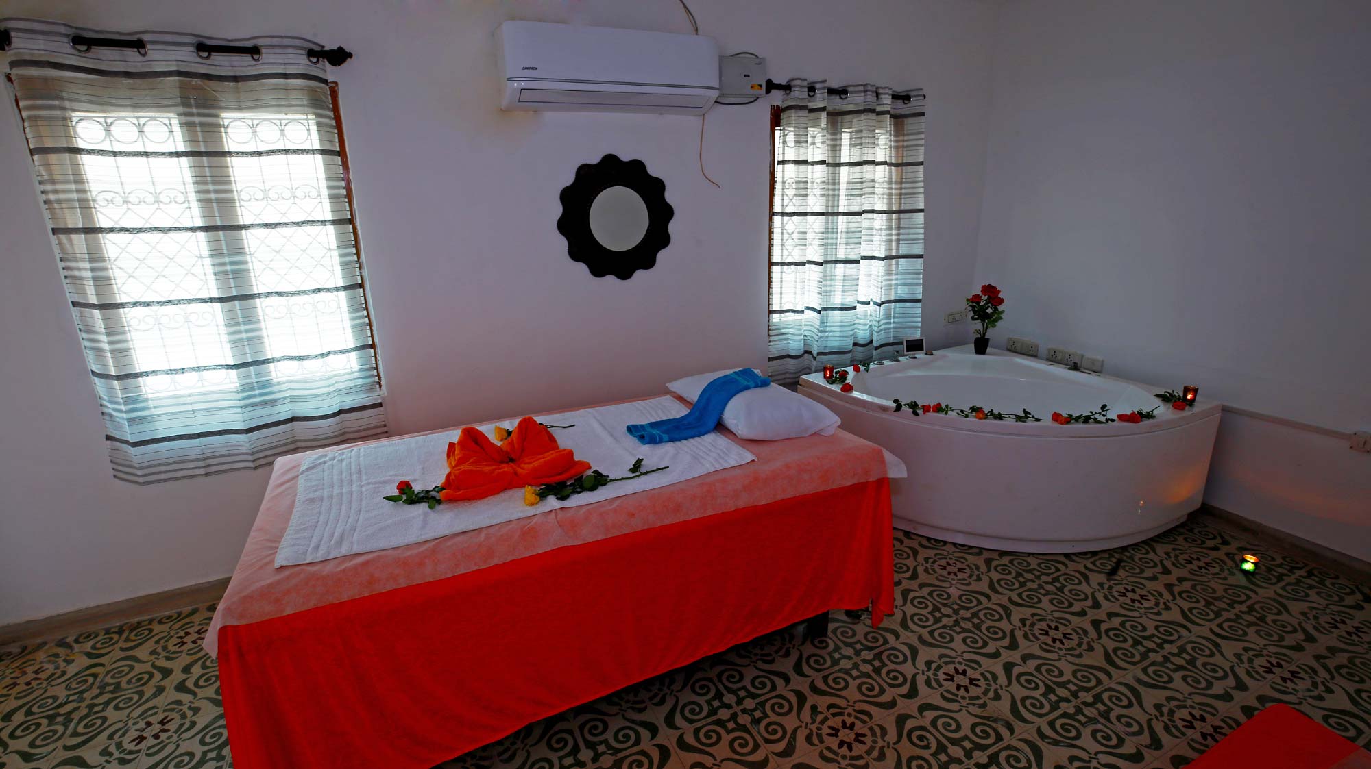 A room in a massage spa features bath tub, massage table with pillow, towel and flowers laid down, Air Conditioner, two windows with curtains and a mirror.