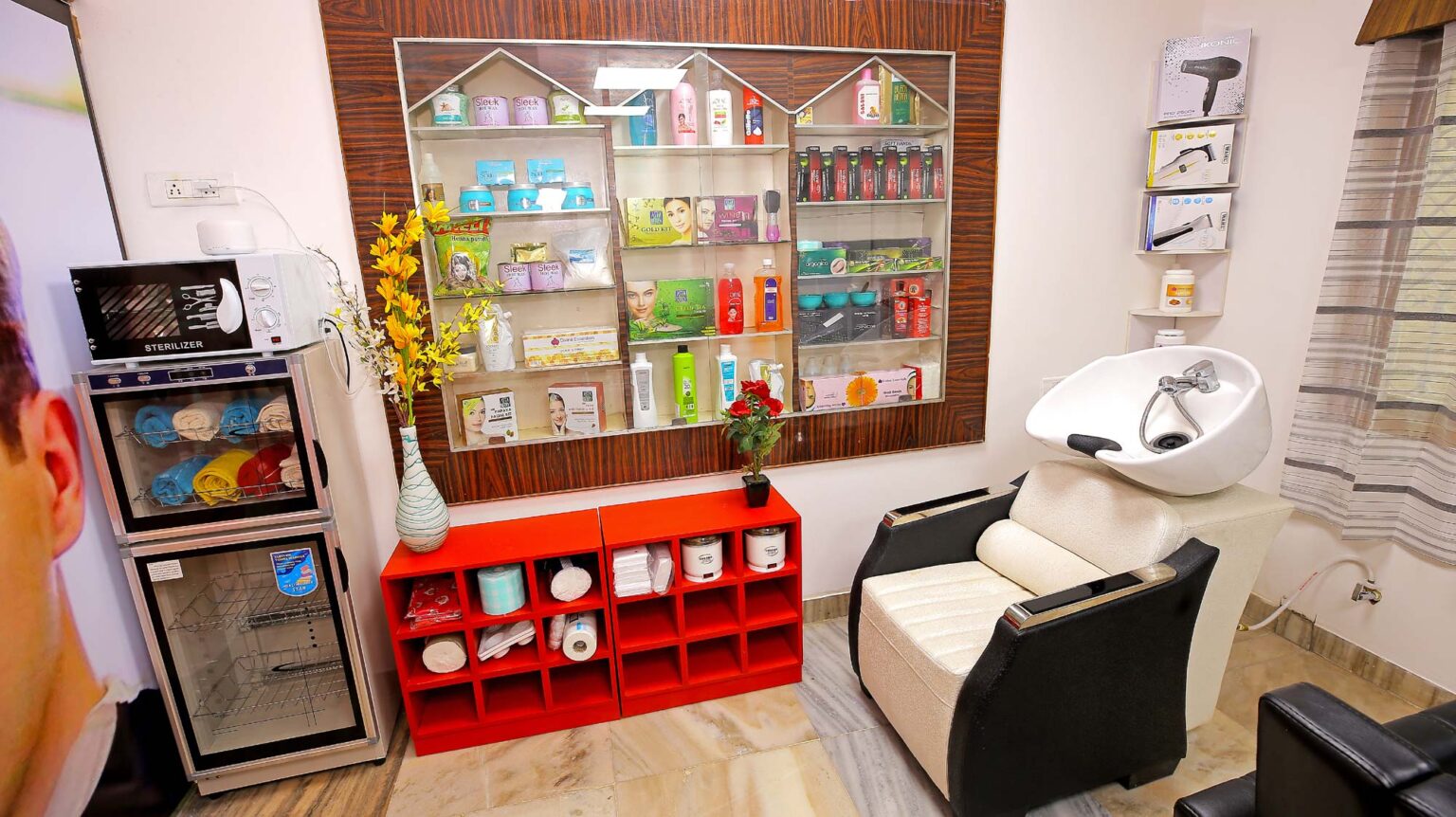 A hair care salon features various products inside a glass shelf, colourful towels, two flower pots, a couch with a hair dryer and three hair dryer products kept on the wall.