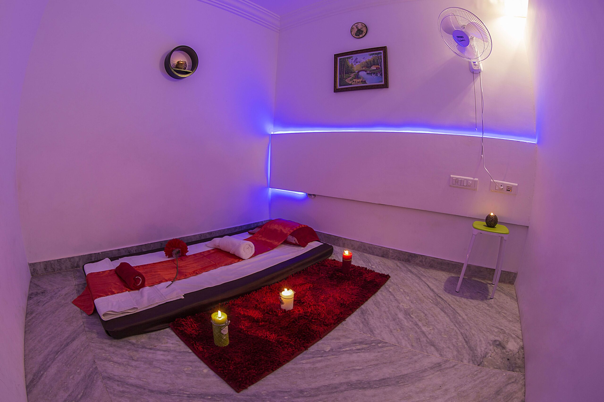 A longer view of relaxed massage room in a spa with a flat bed and towels rolled on into it . A beautiful red color rug with different colored big candles lighten up . A wall mounted fan to provide fresh air in the room. Some candles are kept on the chair and on the wall to improve the freshness of the room with mild lighting in the background.