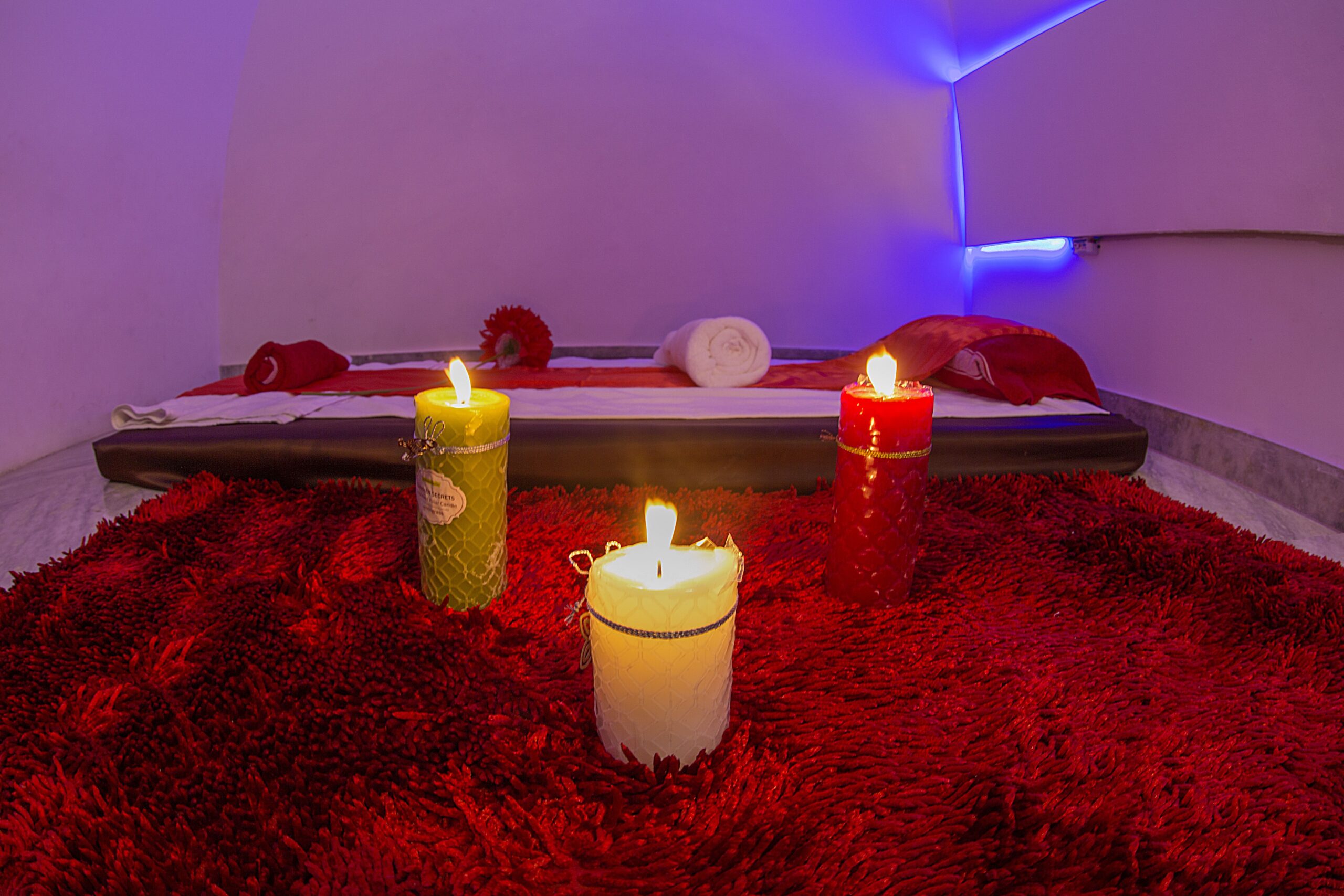 A closer view of a relaxed massage room in a spa with a flat bed and towels rolled on into it A beautiful red colour rug with different coloured big candles lighten up. A mild light in the background gives a pleasant environment.