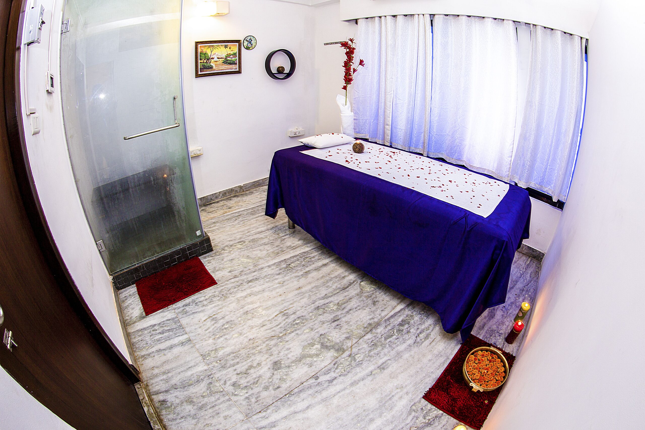 There was a glass cabin in a corner of the room, two lighted candles on the floor, a water container with flower petals in it, and a lamp. The massage table is covered in a blue satin cloth along with candles and flower petals and has a white pillow and white linens on top.