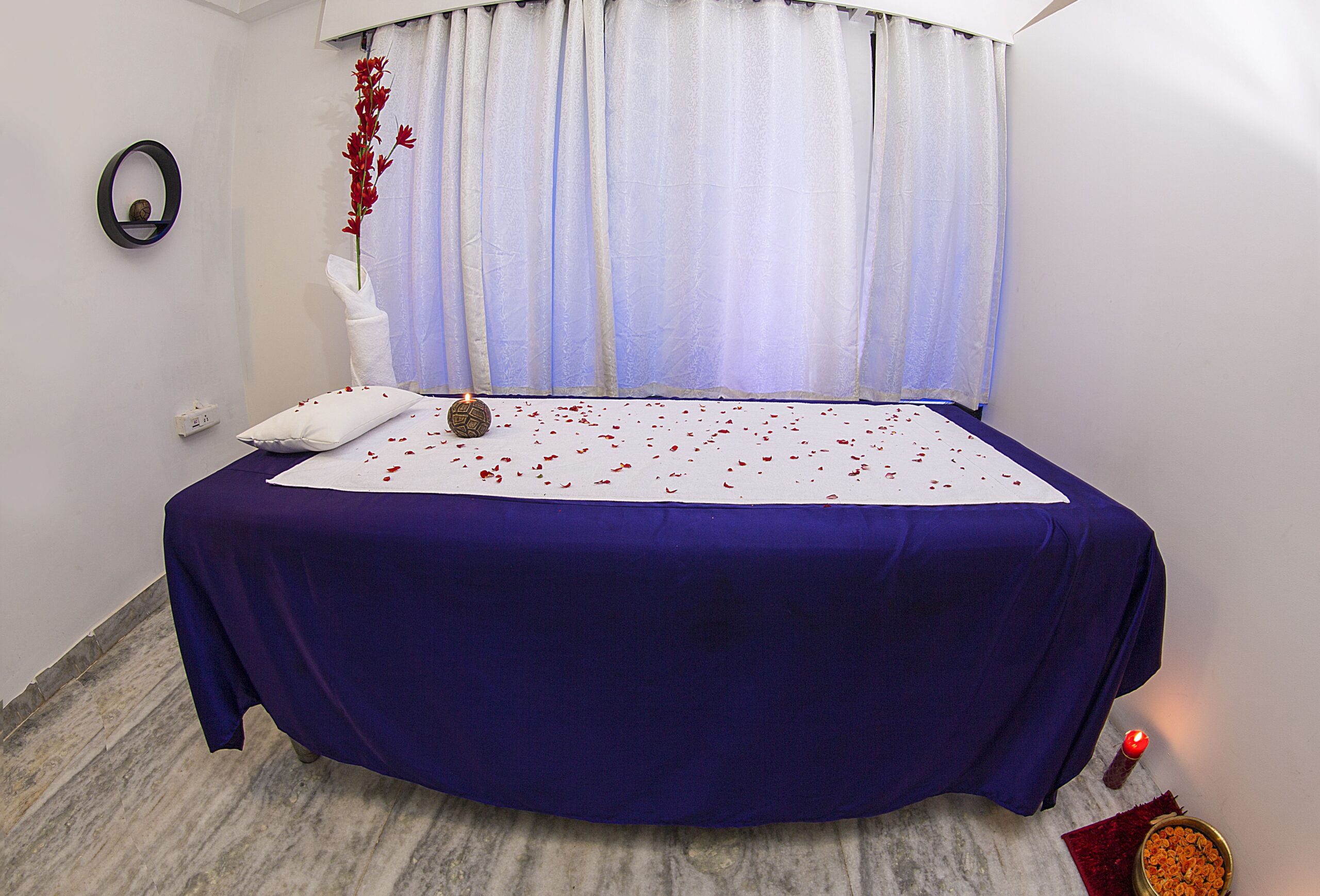 Side view of the beautifully decorated bed with flowers. A relaxed massage room in a spa with a flat bed and towels rolled on into it. Aromatic candles and fresh flowers are placed for freshness in the room.
