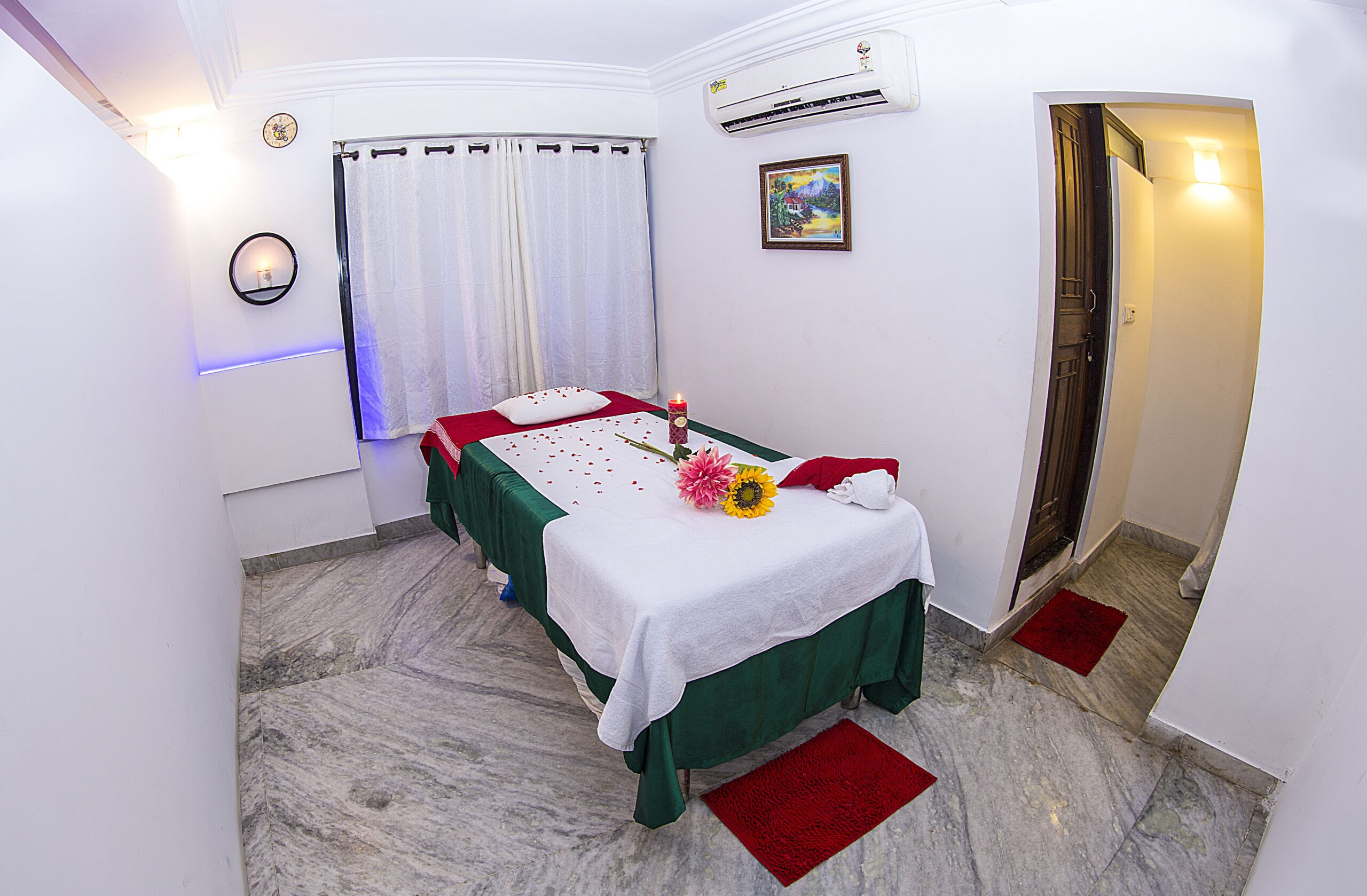The full view of the air-conditioned room in the saloon with a comfortable bed, pillow and towels rolled into it. The room is lightened up with mild light arrangements and neat curtains. Aromatic candles are lit up on the bed and the wall for the freshness in the room. Washroom facilities are also attached for customers comfort.