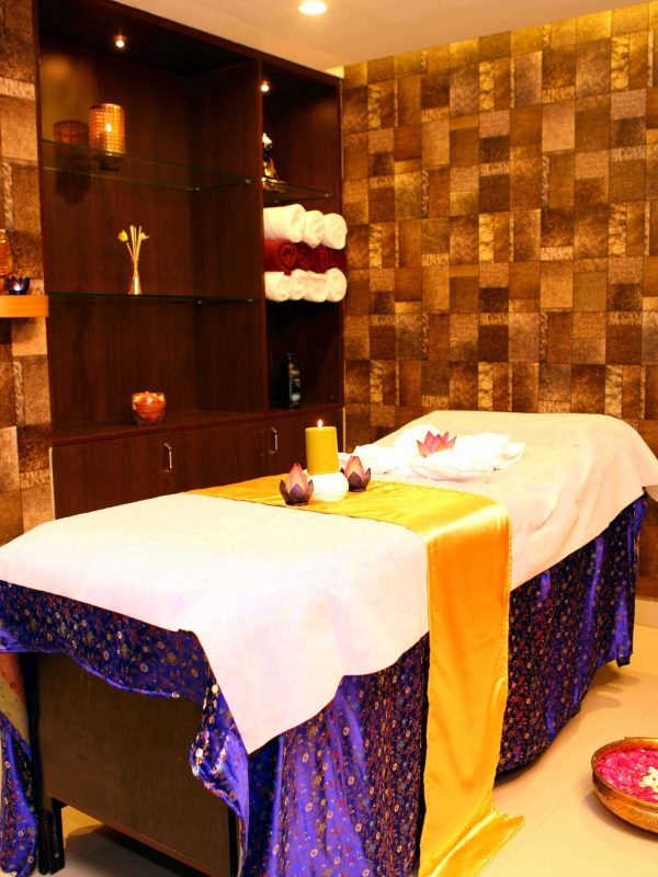 Inside view of spa & massage room depicting an ultra-soft blue satin, cloth covers the run through the bed, topped with a white bedcover. Yellow satin cloth adorns the centre with a yellow candle and two lotus flowers. A pink lotus lies on top of a white towel. A cupboard is attached to the wall with some nice accessories