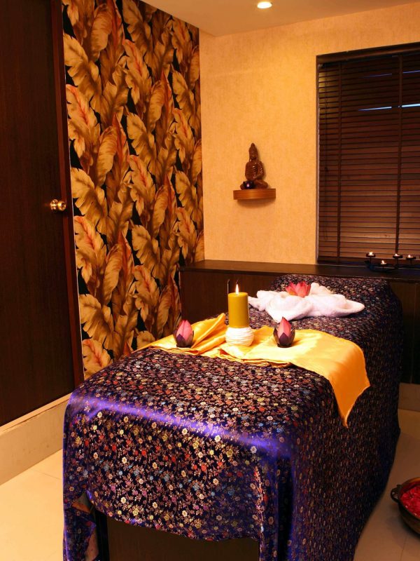 Spa towels, burning candles and lotus flowers are put on the massage table in the massage centre, featuring leaf designed wall, wood buddha idol and spacious window