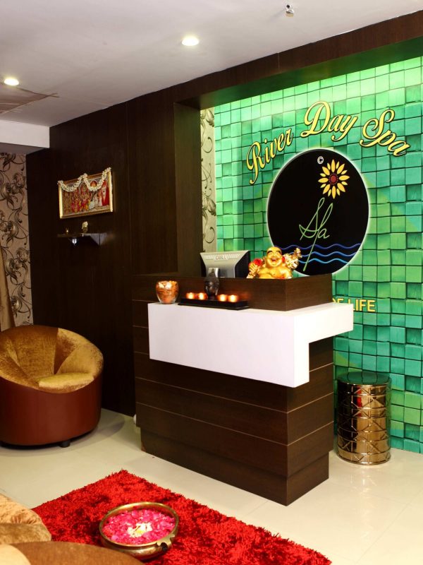 A reception area of a professional spa salon decorated with bright lighting, candles, flowers while its wall bearing the logo of River day spa. The room features couches to sit in and a Hindu god's photo frame