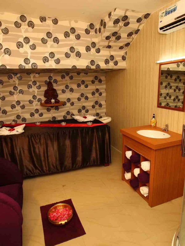 A side view of a fully air-conditioned spa room with a neatly decorated bed with fresh flowers and aromatic candles. The wall is decorated with beautiful wallpaper for an elegant look for the spa. It has a premium sofa's at the side of the bed and wash basin with a rack of towels.