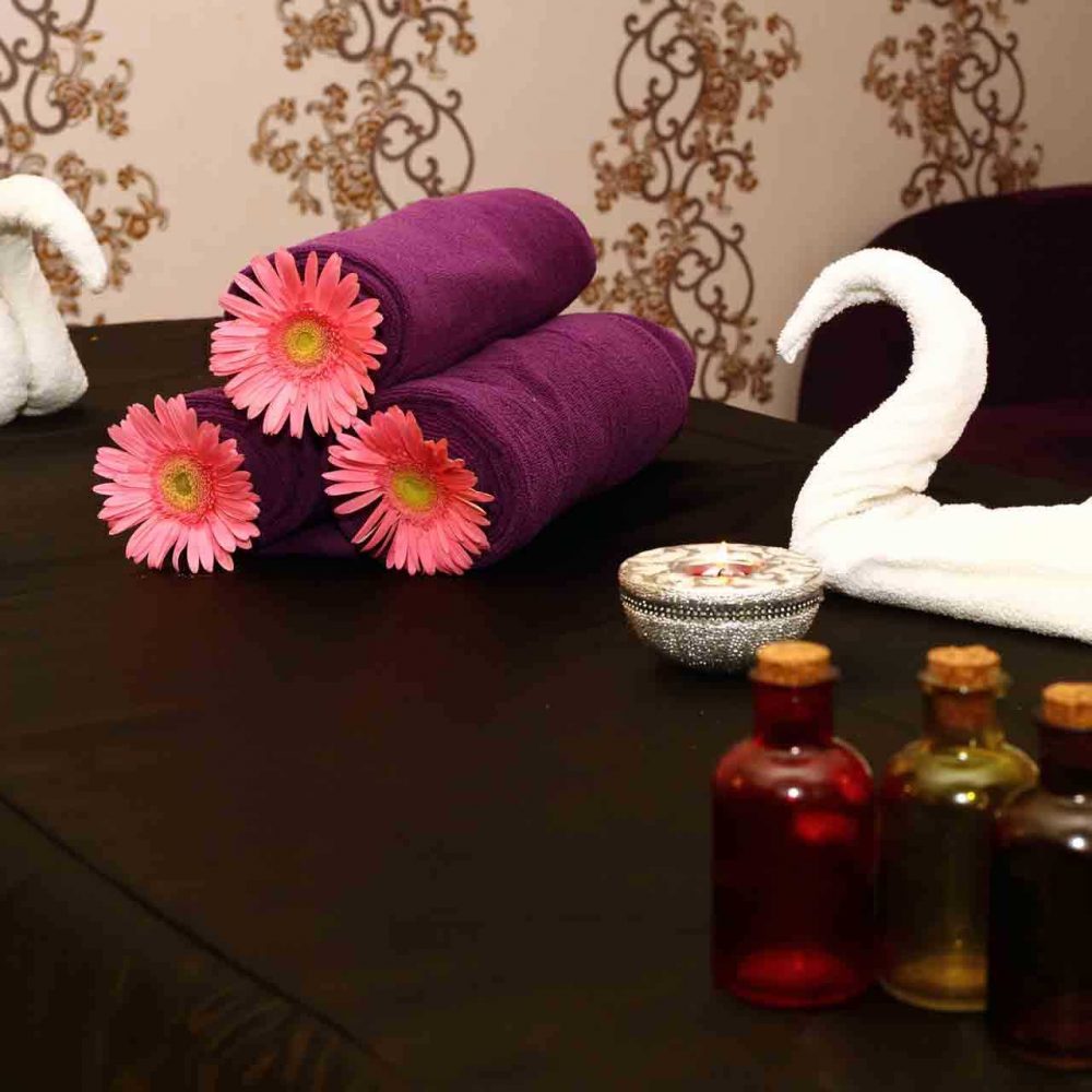 Spa room with a table displayed three magenta-coloured rolled-up towels, each with a gerbera flower in the middle, two white swan-shaped towel kept to the side, and three different perfumes bottles